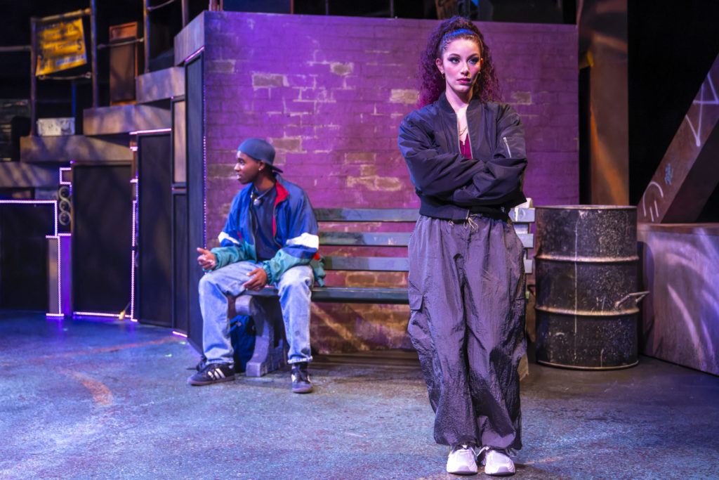 A black man sits on a bench in front of an exposed brick wall. A wall is standing in front off to the side with her back to the man and her arms crossed.