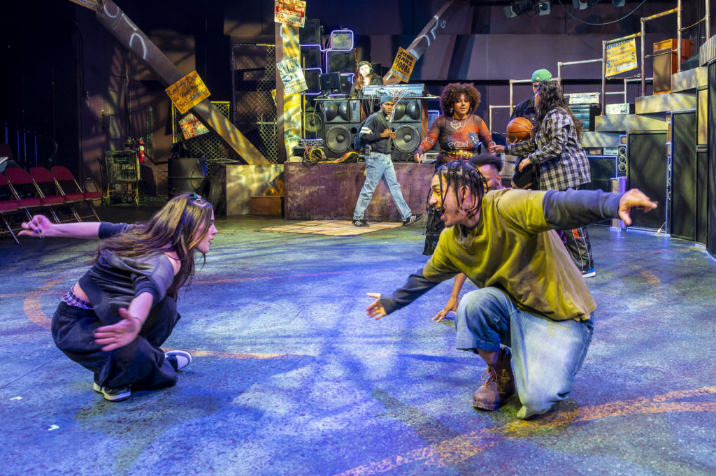 A white woman on the left and a Black man on the right are on their knees facing up, presumably during a rap battle