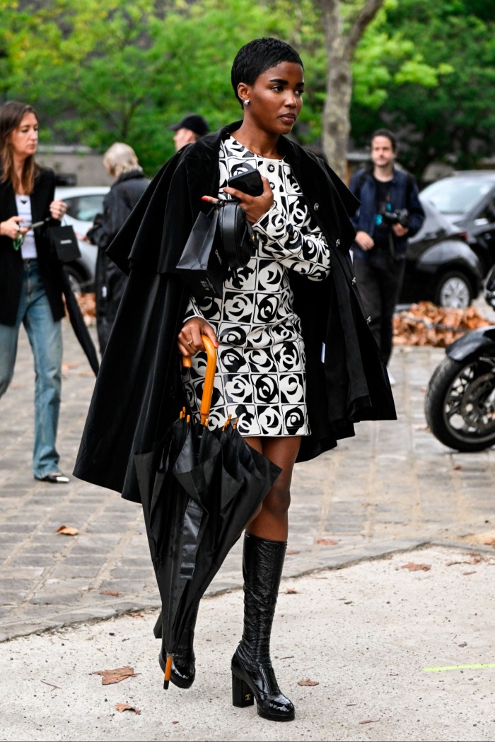 A woman walks on a street wearing a black and white mini dress and boots, with a black coat slung over her shoulders