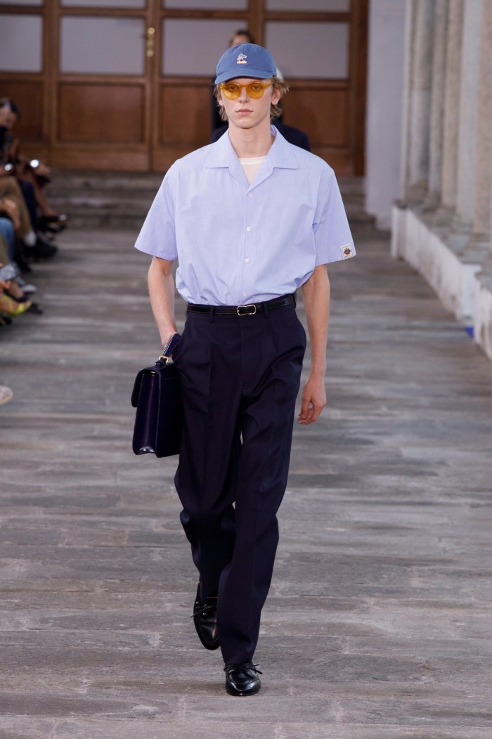 male model on runway wearing a purple shirt, black trousers and shoes and a baseball cap