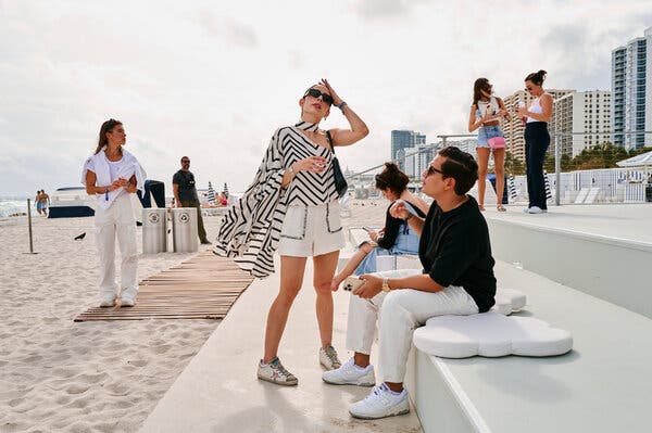 A woman, center, standing on a crowded beach. She has one hand held to her forehead and is wearing sunglasses, a one-shoulder white top with zigzagging black stripes, white shorts with black stitching around the pockets and white sneakers.