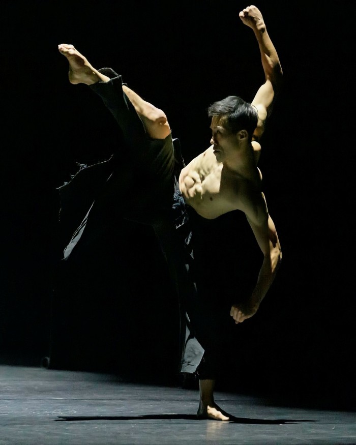 A shirtless man in black trousers bends back as he lifts his right left high