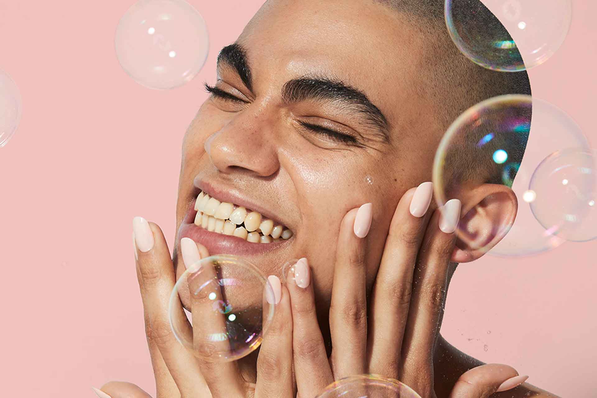 Model smiling against pink background, wearing light pink press-on nails.