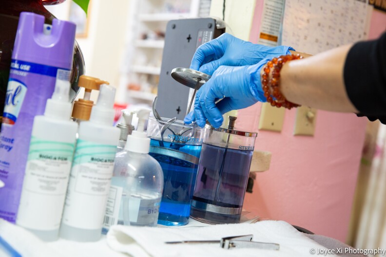 Two blue gloved hands take a metal instrument out of a container filled with blue-colored disinfectant. 