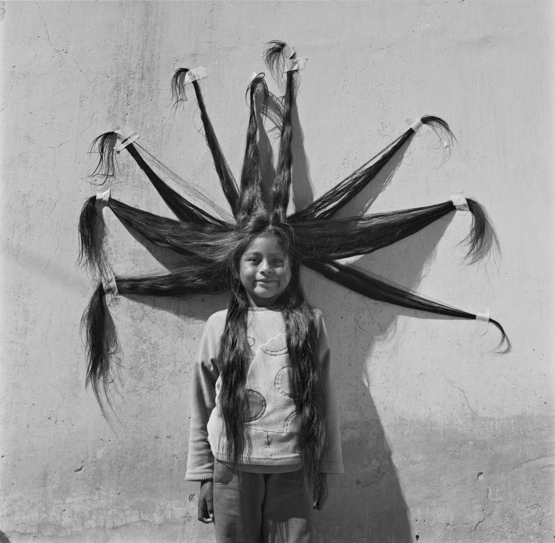 a black and white photo of a young girl whose hair is taped to a wall in a star-like shape smiling for the camera