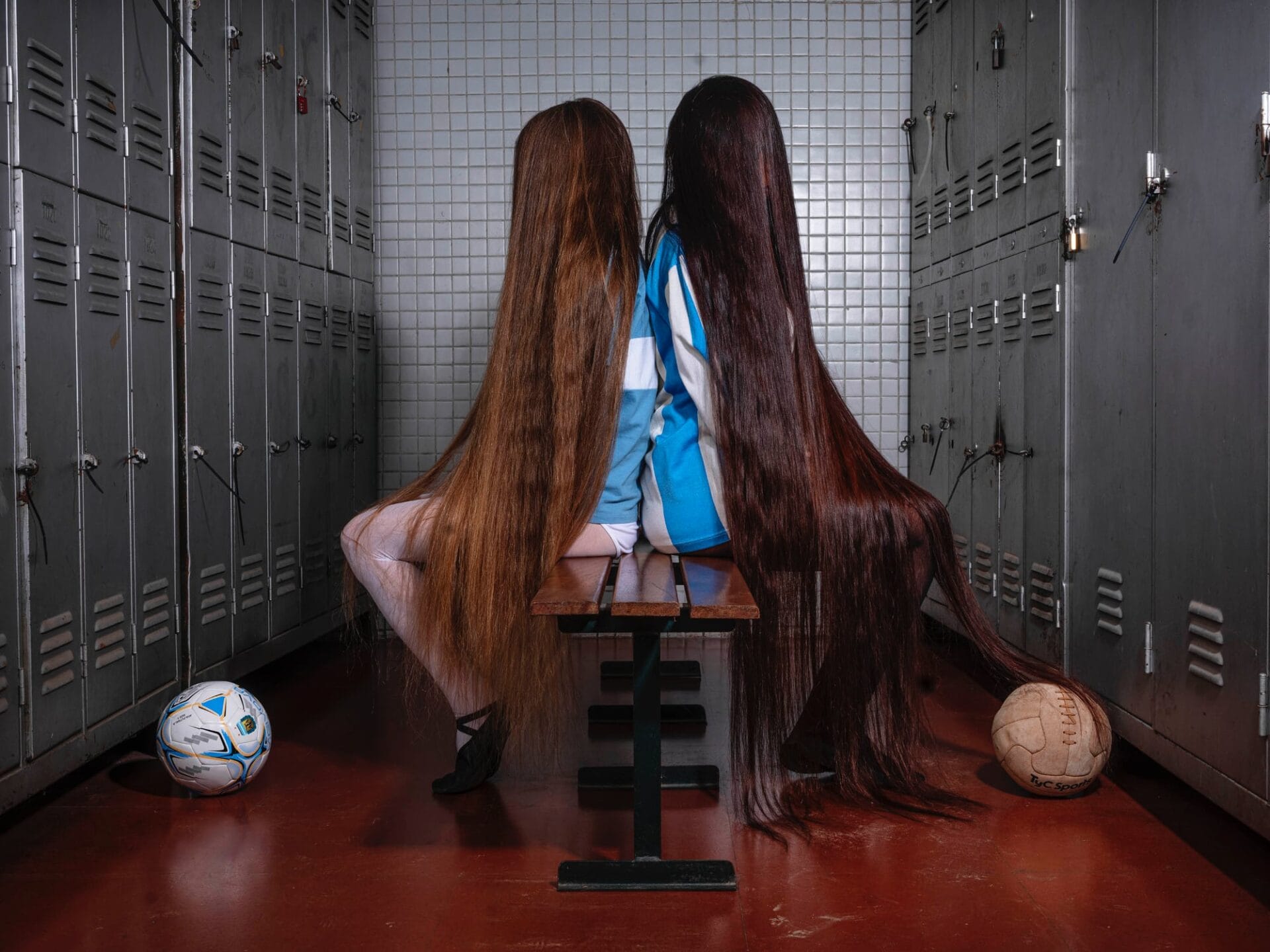 two young girls masked by their hair sit back to back on a locker room bench