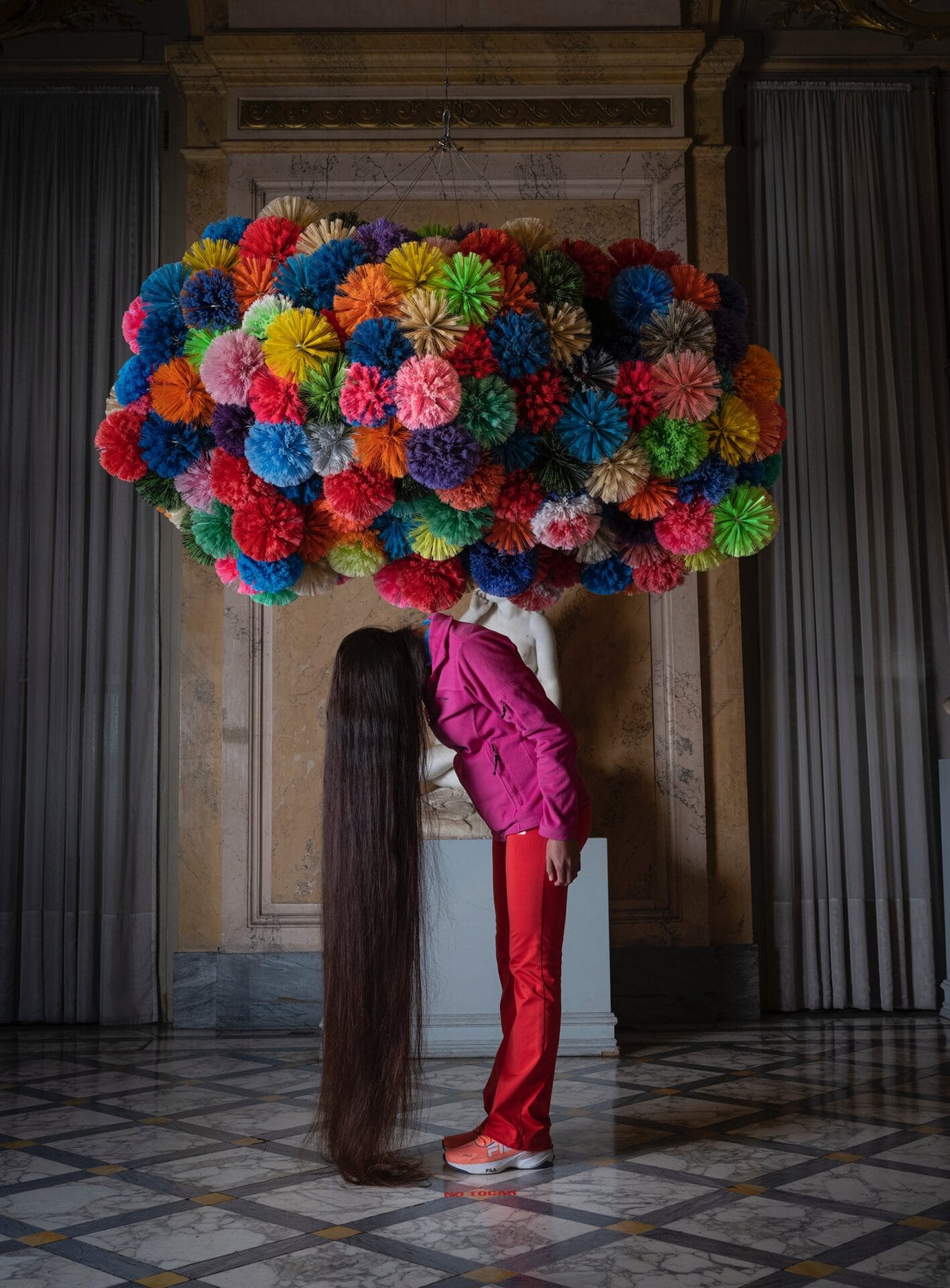 a woman stands bent over with her hair pooling on the floor. a colorful puffy installation is above her