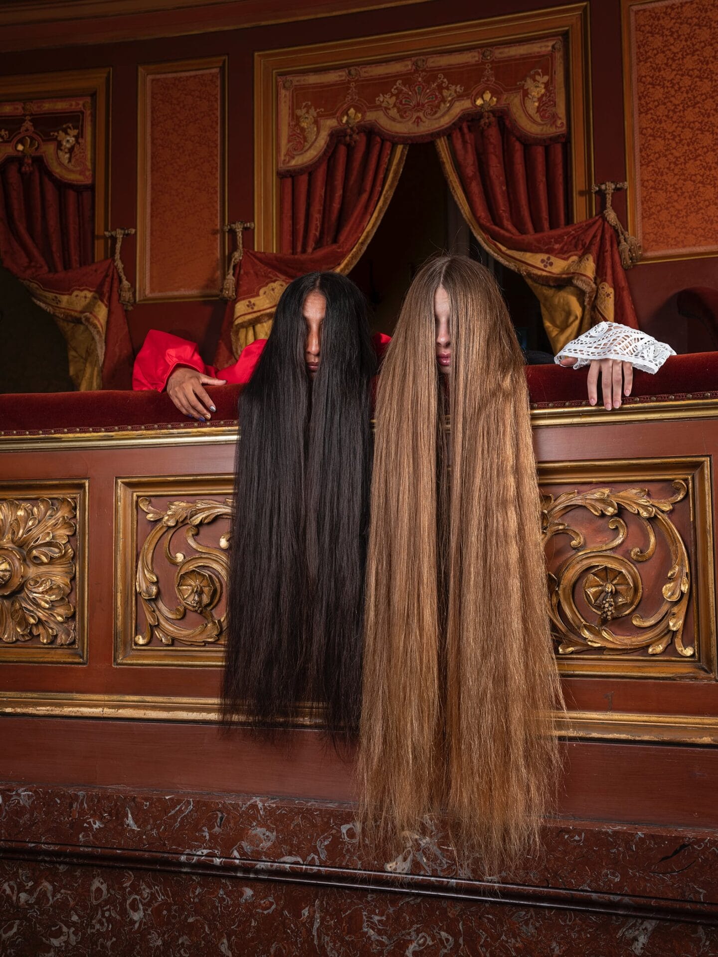 two women rest their chins on an ornate theater balcony with their hair hanging down