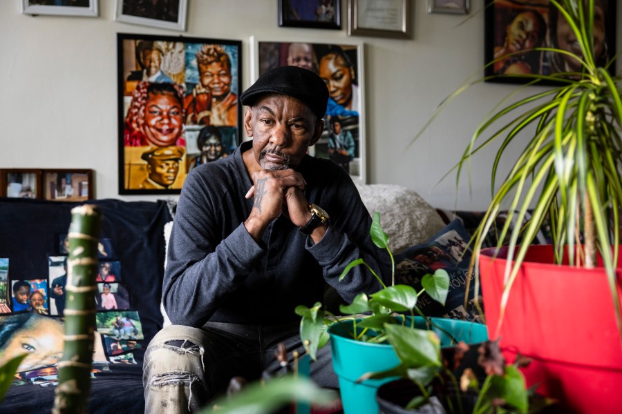 Arther Sanders sitting on living room couch with plants on table in front of him and paintings and photos hanging on wall behind him