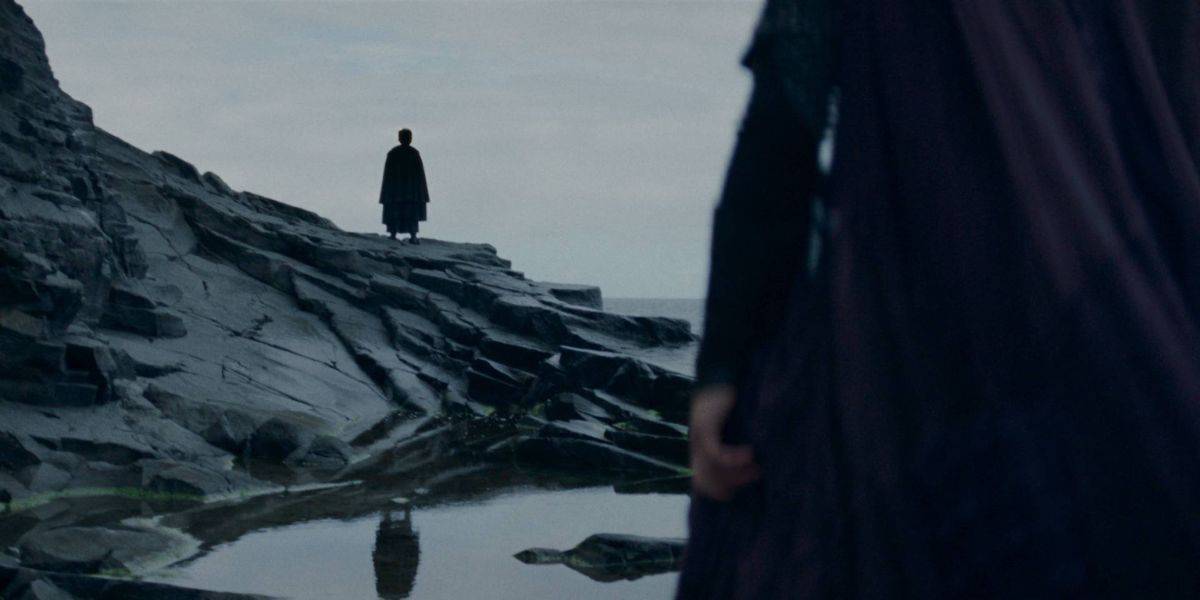 An acolyte stands silhouetted at the edge of a rocky cliff by the ocean, facing away. A second person, partially blurred and closer to the foreground, looks towards the figure on the cliff. The scene has a dramatic, somber feel with a muted color palette.