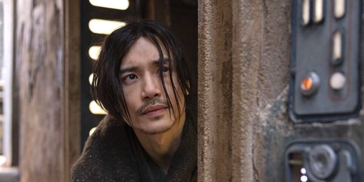 A man with long, dark hair and a mustache peers out from behind a weathered wall. An acolyte of forgotten lore, he has a contemplative expression. The surroundings appear rustic and dimly lit, featuring an industrial control panel on the right side of the image.