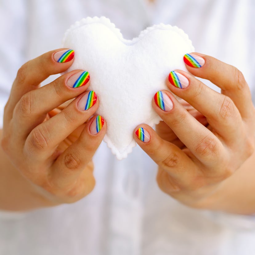 Hands with Pride nails sporting rainbow stripes holding a white heart-shaped pillow.