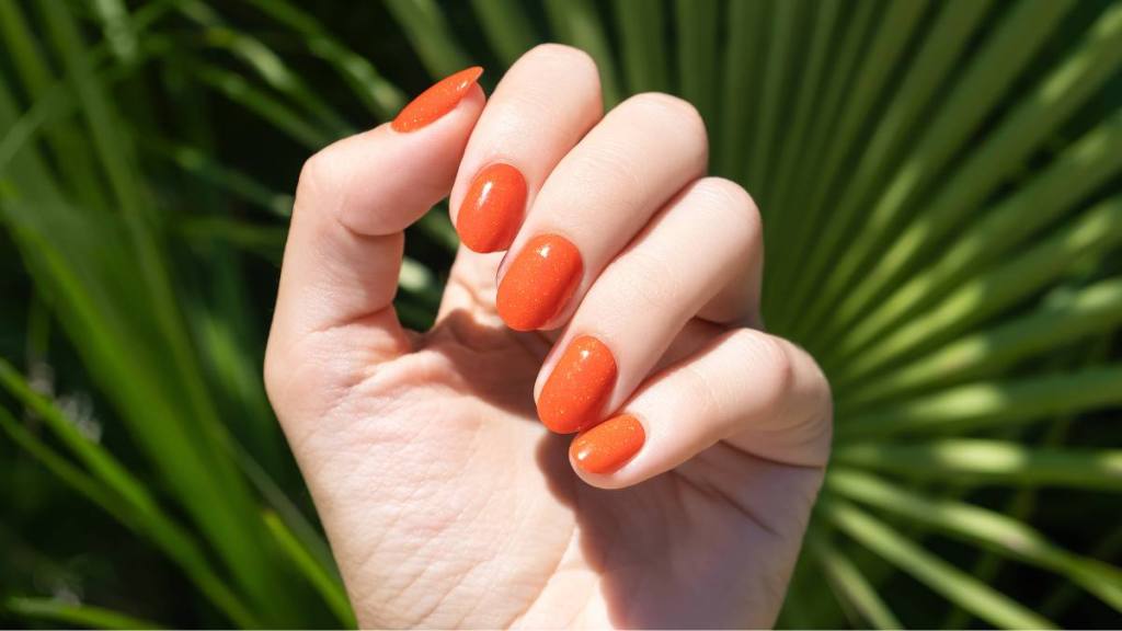 Female hand with orange nail design. Orange nail polish manicure. Female hand on a tropic plant leaf background