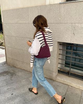 Woman wearing white button-down shirt, striped sweater, light-wash jeans, oxblood bag, and slip-on backless loafers.