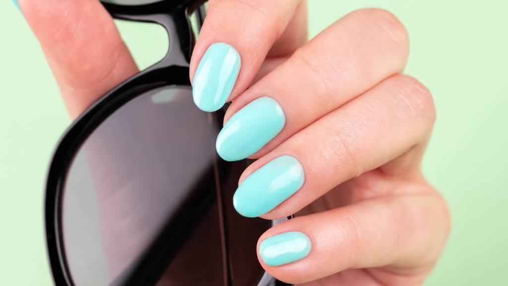 Female hand with beautiful manicure - mint blue nails with sunglasses on green background, closeup, selective focus