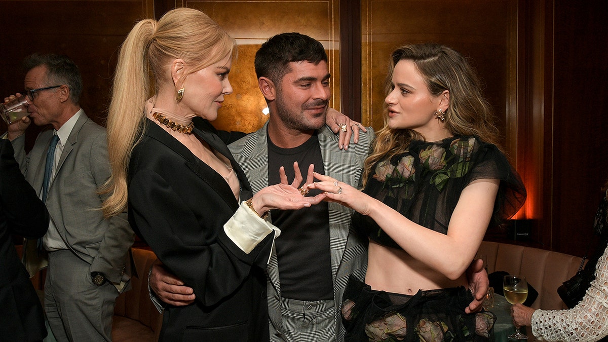 Nicole Kidman in a black blazer speaks with Joey King in a lacey black two piece set as they both have their arms wrapped around Zac Efron in a black shirt and grey suit