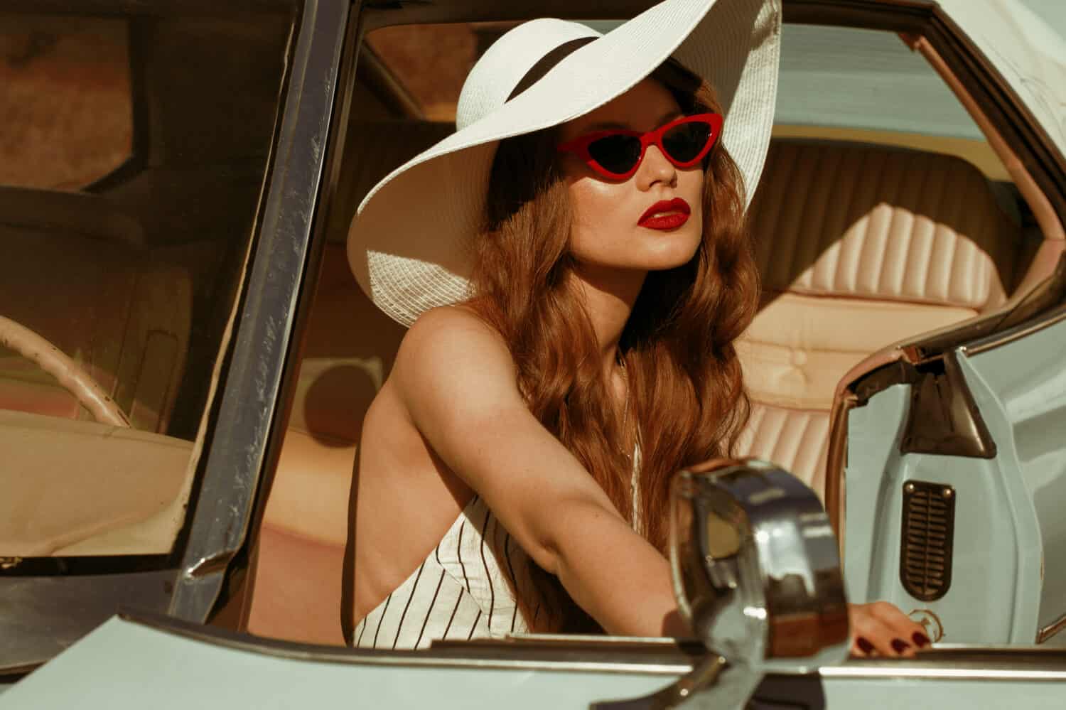 Street fashion portrait of young elegant luxury lady wearing white sunglasses, wide brim hat, striped linen jumpsuit, posing in the retro car. Copy, empty space for text