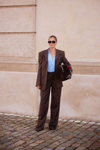 A woman wearing an oversized brown blazer with a blue button-down shirt, brown tailored pants, sunglasses, and a brown suede clutch.