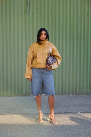 A woman wearing a mustard-color leather jacket, denim bermuda shorts, Jacquemus heels, and a Loewe bag with gold bag charms.