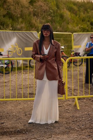 A woman wearing a brown leather blazer with a white knit maxi dress, white heels, a brown bag, and large sunglasses.