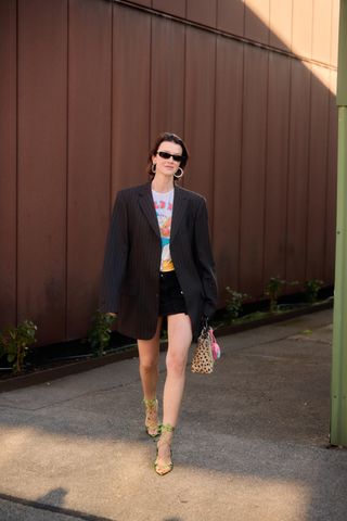 A woman wearing a pinstripe oversized blazer, a white graphic t-shirt, a black denim miniskirt, a leopard bag, and green lace-up heels.