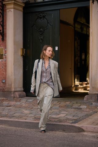 A woman wearing a pale green oversized blazer with matching trousers, black heels, and a striped button-down shirt.