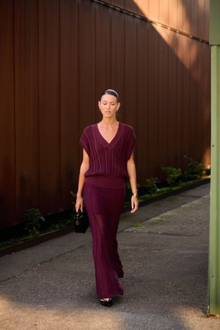 A woman wearing a burgundy sweater vest, burgundy sheer maxi skirt, black heels, and a micro black bag.