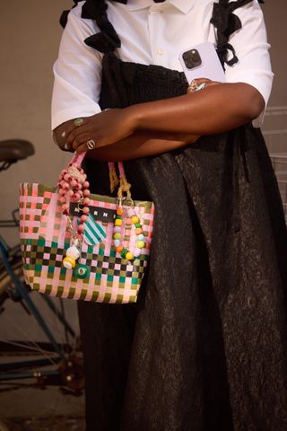 A woman wearing a button down shirt under a black dress, carrying a woven Marni colorful tote bag with bag charms.