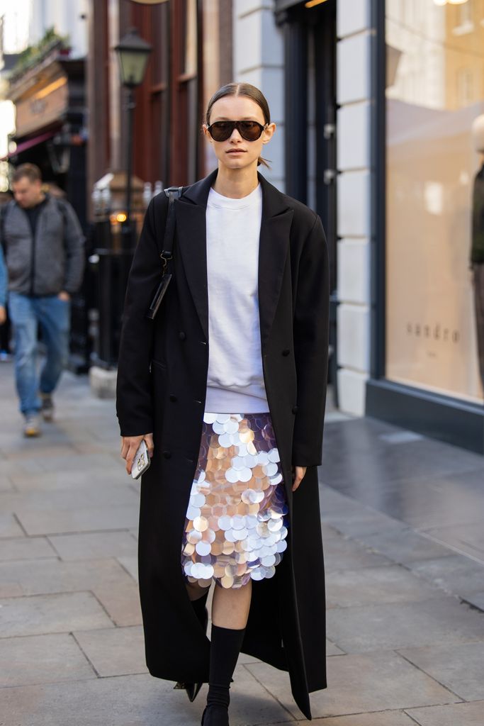 A guest wears sequined skirt, black long coat, Balenciaga bag, sunglasses, mid high socks outside Bora Aksu during London Fashion Week September 2024 on September 13, 2024 in London, England.