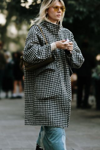 A Milan fashion week guest wearing blue jeans and a black and white houndstooth coat walking on the sidewalk.