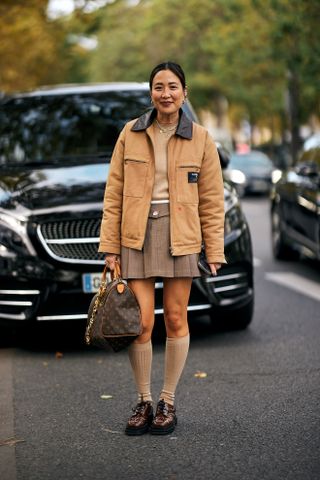 Woman at Fashion Week walking in the street wearing brown loafers, a brown mini skirt, tan socks, a tan barn jacket, and a Louis Vuitton duffel bag.