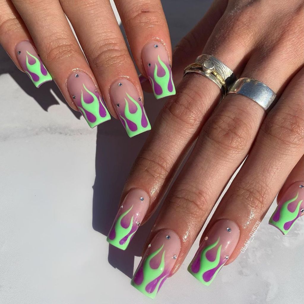 Square nails with clear bases featuring vibrant green and purple flame designs, accentuated with small, subtle rhinestones, creating a bold and edgy look with a touch of sparkle.