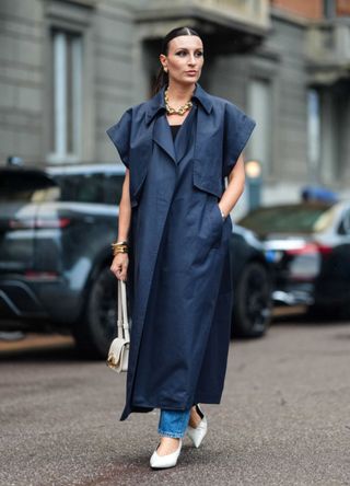 Carlotta Rubaltelli wears gold earrings, a gold necklace, a navy blue wide short-sleeve midi dress, mixed metal bracelets, a white leather bag, light blue denim jean pants, and white pointed-toe heels, outside Onitsuka Tiger, during the Milan Fashion Week Spring/Summer 2024-2025 on September 18, 2024 in Milan, Italy.