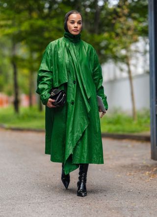 A guest wears bright green oversized Ferragamo leather trench coat, dark brown cap / scarf over the head, shiny black Ferragamo leather bag, shiny black pointed toe heeled leather boots, outside Ferragamo, during the Milan Fashion Week Spring/Summer 2024-2025 on September 21, 2024 in Milan, Italy