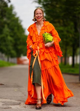 A guest wears a dark green pattern hat, gold earrings with pearls, gold layered necklaces, bright orange sheer mesh ruffled long sleeve open flowy midi dress, dark green wide-legged pants, dark brown open-toe platform heels, outside Ferragamo, during the Milan Fashion Week Spring/Summer 2024-2025 on September 21, 2024, in Milan, Italy