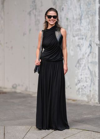 Sophia Roe wears black sunglasses, silver earrings, black sleeveless with shiny black detail Ferragamo maxi dress, black leather bag, outside Ferragamo, during the Milan Fashion Week Spring/Summer 2024-2025 on September 21, 2024 in Milan, Italy