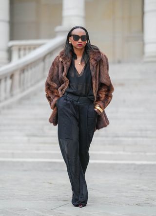 Emilie Joseph wears black sunglasses, gold and red earrings, a brown faux fur jacket, a gold necklace, a black low-neck top with transparent details, chocolate and golden bangles, black wide-leg pants with transparent details, burgundy Lee Patent Leather pointed-toe from Saint Laurent, during a street style fashion photo session in Paris, France