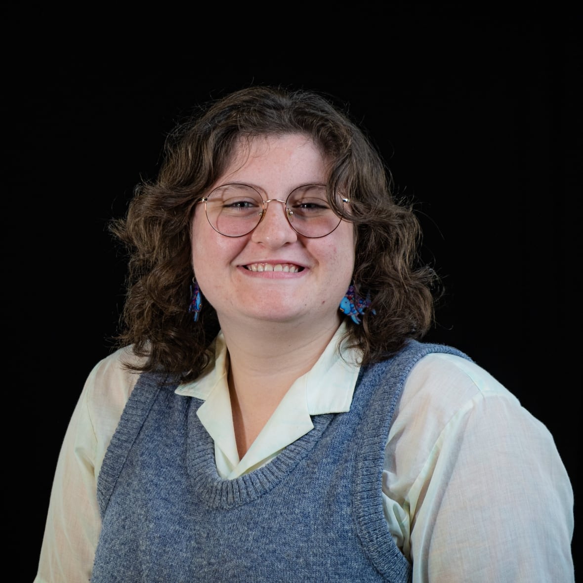 Portrait of young, Caucasian woman with curly brown, shoulder length hair and wire-framed glasses. They wear a light blue knitted vest over a white-collared shirt. They are wearing blue, dangly earrings. 