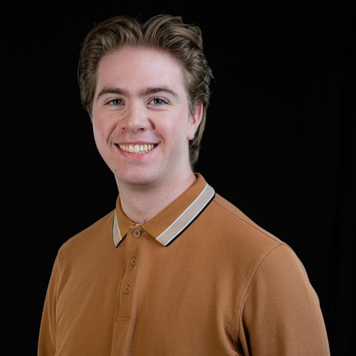 Young, Caucasian man with short, dirty blond hair smiles with his teeth showing. He is wearing a mustard top with a collar which is buttoned up. 