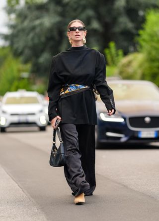 A guest wears black sunglasses, gold earrings, black oversized blazer jacket backwards, black blue pattern/print scarf belt, shiny leather bag, black loose pants, outside Jil Sander, during the Milan Fashion Week Spring/Summer 2024-2025 on September 18, 2024 in Milan, Italy