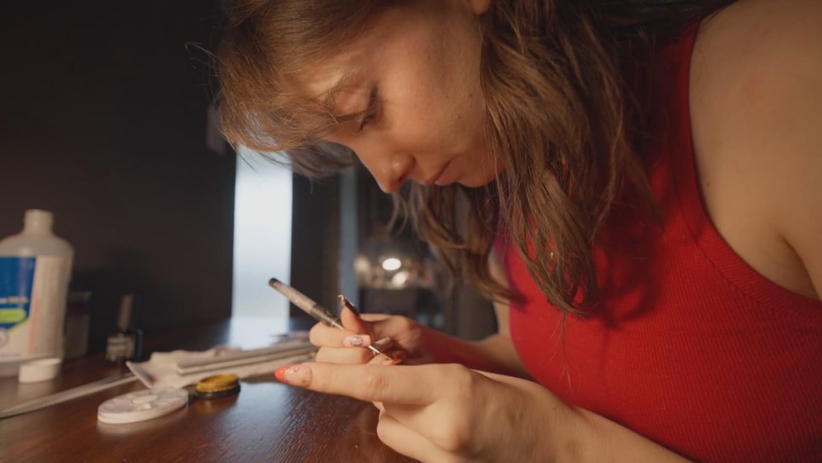 Young woman with shoulder length light brown hair leans over a table painting a design on a fake fingernail. She wears a red tank top. 