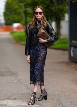 A guest wears black sunglasses, a shiny black leather jacket, a black sheer mesh floral dark blue pattern midi skirt, black sheer mesh tied up open toe heels, a dark brown Ferragamo leather bag, and a silver watch, outside Ferragamo, during the Milan Fashion Week Spring/Summer 2024-2025 on September 21, 2024, in Milan, Italy.