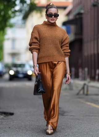 Tatyana Kodzayeva wears gold hair accessories, black sunglasses, gold earrings, a dark brown oversized turtleneck sweater, a shiny dark brown copper flowy satin skirt, gold bracelets, a shiny black leather bag, shiny gold pointed-toe strappy heels, outside Alberta Ferretti, during the Milan Fashion Week Spring/Summer 2024-2025 on September 2024 in Milan, Italy.