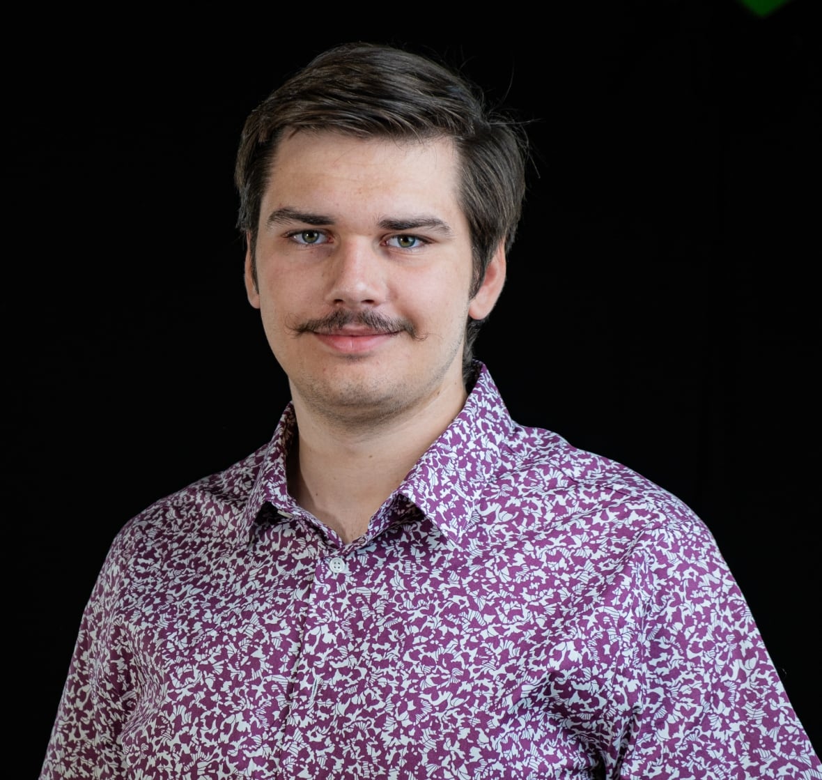 Portrait of young, Caucasian man with short dark hair and a short moustache. He is wearing a purple, flower-patterned button down shirt with a collar. 