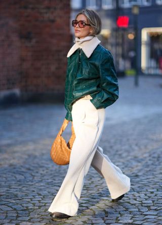 A guest wears sunglasses, a green leather jacket with white fluffy collar, white flared pants palazzo pants , a brown woven leather bag, outside Herskind, during the Copenhagen Fashion Week AW24 on January 31, 2024 in Copenhagen, Denmark.