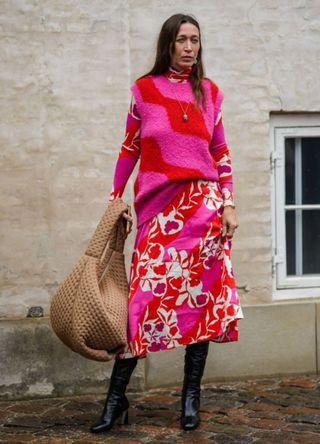 Alana Hadid wears neon red and neon pink wool sleeveless pullover, a white with red / neon pink print pattern long sleeves midi dress, a beige matte leather quilted oversized handbag, black shiny leather heels knees boots / high boots , outside Marimekko, during the Copenhagen Fashion Week Spring/Summer 2024 on August 09, 2023 in Copenhagen, Denmark.