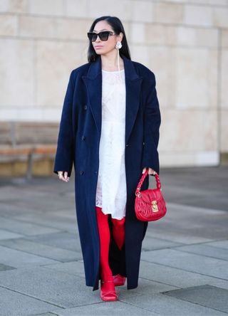 Miki Cheung wears Dior sunglasses, flower shaped earrings, a white dress with floral embroidery, a navy blue long coat, red leggings, a red leather Miu Miu bag, red leather shoes, outside OperaSport, during the Copenhagen Fashion Week AW24 on January 31, 2024 in Copenhagen, Denmark