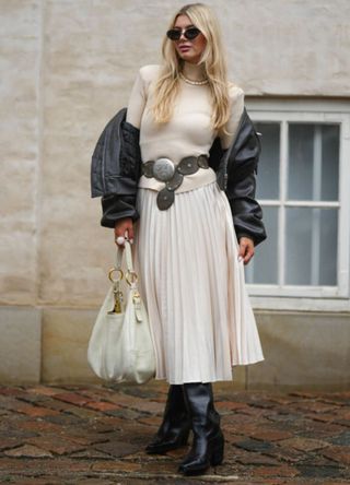 A guest wears black sunglasses, a beige ribbed wool turtleneck pullover, a white pearls necklace, a black shiny leather bomber coat, a dark gray matte leather western belt, a white latte pleated / accordion midi skirt, a white latte matte leather handbag, black shiny leather pointed / block heels ankle boots , outside Marimekko, during the Copenhagen Fashion Week Spring/Summer 2024 on August 09, 2023 in Copenhagen, Denmark.