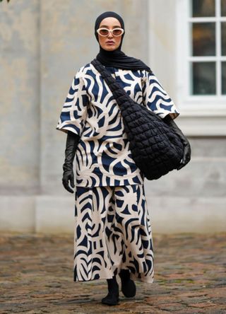 A guest wears a black scarf as a headband, white sunglasses, a navy blue and white latte print pattern oversized t-shirt, matching navy blue and white latte print pattern oversized flowing pants, a black fabric quilted oversized shoulder bag, black shiny leather high gloves, black socks, black shoes , outside Marimekko, during the Copenhagen Fashion Week Spring/Summer 2024 on August 09, 2023 in Copenhagen, Denmark.