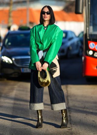 A guest wears black sunglasses, a black pearls with white heart pendant necklace, a green t-shirt, a green blazer jacket, a beige / black / green print pattern wool pullover, black rolled-up denim pants, a gold shiny varnished leather handbag from Coperni, gold shiny leather pointed heels ankle boots , outside Munthe, during the Copenhagen Fashion Week Autumn/Winter 2023 on February 02, 2023 in Copenhagen, Denmark.